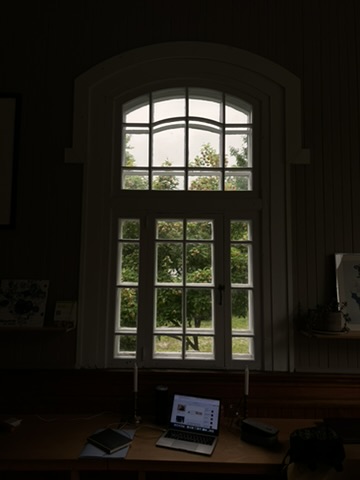 A tall church window above a desk with a laptop and candles.